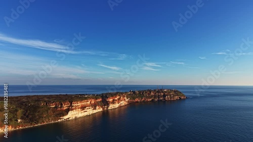 Wallpaper Mural A coastal cliff juts out into the sea under a blue sky. The water is calm, and some trees and structures are visible along the cliff edge. This scene shows nature and human presence together. Torontodigital.ca