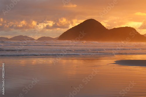 Golden sunrise in Riviera beach, Bertioga, Brazil. Light reflections on sky and water, mountains shilouetttes and cloudy sky. Waves breaking on water edge.