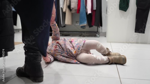 Young child in store throws tantrum on floor, upset and crying while lying down amidst clothing racks.
