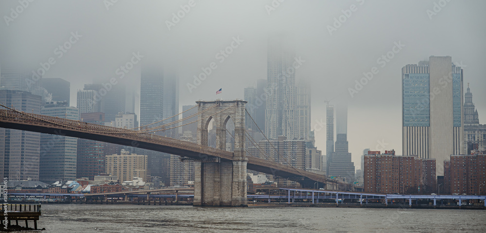 Obraz premium Fog covers Brooklyn Bridge and Manhattan skyline on a cloudy day. Fog fills the air over Brooklyn Bridge and the Manhattan skyline in New York City during an overcast moment.