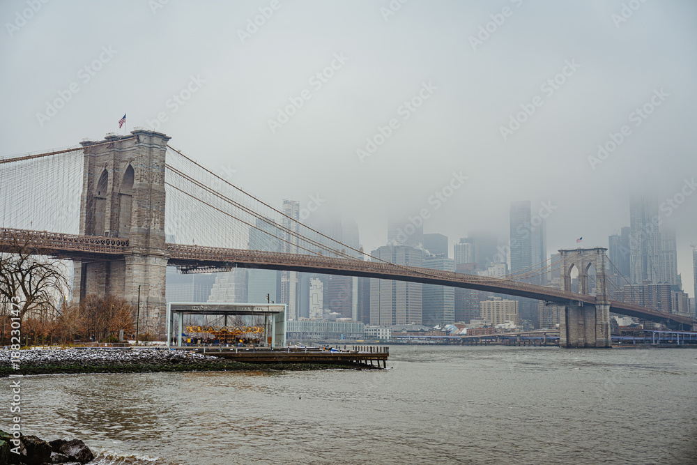 Fototapeta premium Brooklyn Bridge in fog with city skyline and waterfront view. Brooklyn Bridge stands in fog with a view of tall buildings and water.