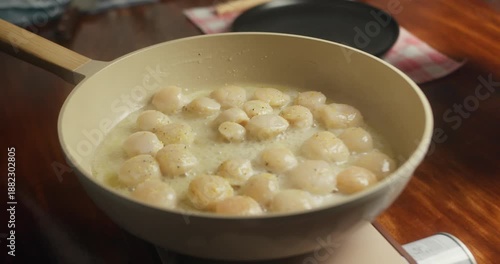 Woman cooking scallops at home in her kitchen, frying sea scallops in a pan on a portable gas stove. Close-up of scallops being turned to achieve an even golden sear, homemade seafood cooking process