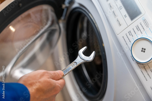 Hand holding a wrench is positioned near the door of a washing machine, showcasing the appliance's control panel and interior during a maintenance task in a home setting