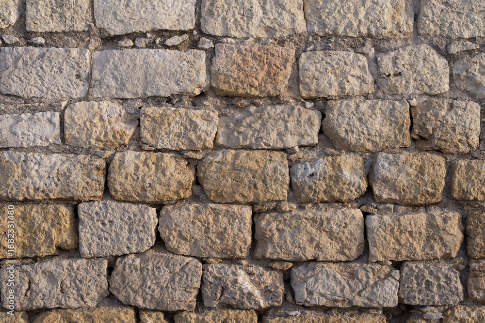 Obraz premium Detailed texture of a weathered stone wall made of rough limestone blocks. The surface shows natural erosion, cracks, and warm tones, ideal for architectural, historical, or design backgrounds.