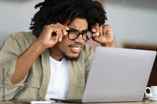 Photos Concentrated young black curly guy in glasses squints and looks at laptop at table with cup in cafe interior
