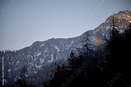 Evening light brushes a rugged alpine ridge above Ruhpolding, Bavaria. Patchy snow clings to the rocky slope between dark spruce and larch, while a clear winter sky fades toward dusk. 