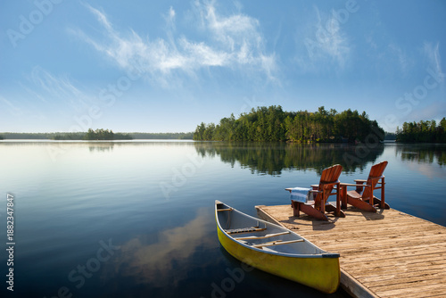 Two wooden Adirondack chairs on a weathered dock in Muskoka, ON, with a blue towel. A yellow canoe rests on the mirror-like lake, with green islands and a clear blue sky reflecting calm cottage life