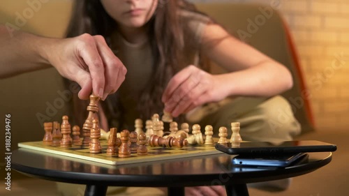Digital detox, playing chess at home. Young girl and her father carefully placing wooden chess pieces on the board, cozy living room, smartphones are lying on the table