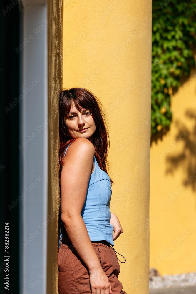 Fototapeta premium Young woman with brown hair is leaning against a yellow wall, showcasing a relaxed pose in a vibrant outdoor setting, capturing a moment of tranquility and warmth
