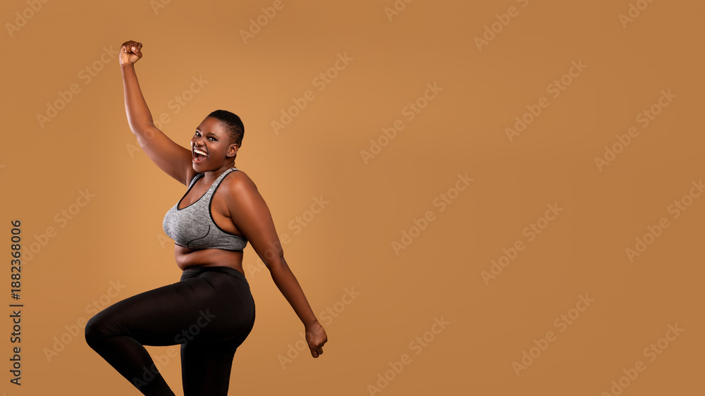 Obraz premium Excited black chubby woman in sportswear dancing, emotional happy lady celebrating success and victory, jumping, cheering and raising clenched fist up, brown studio background, banner, free copy space