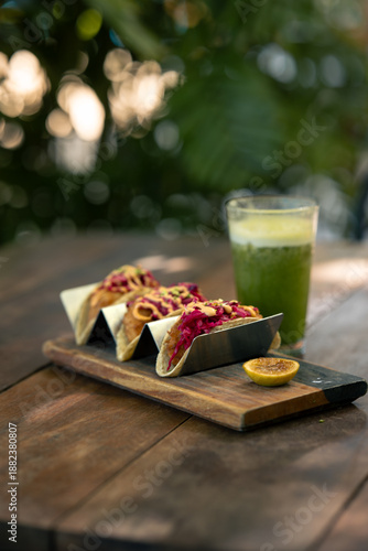 Tres tacos de pollo con betabel servidos sobre una mesa de madera, y un jugo verde con la textura de los árboles al fondo