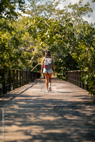 Mujer, hermosa latina y viajera, caminando con su mochila sobre un muelle rodeado de árboles frondosos bajo la luz del sol al atardecer