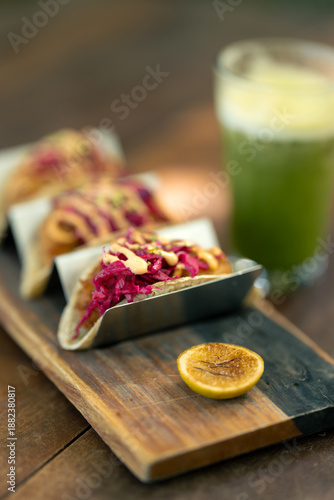 Close up de tres tacos de pollo con betabel servidos sobre una mesa de madera, y un jugo verde con la luz del sol filtrándose entre las hojas de los árboles