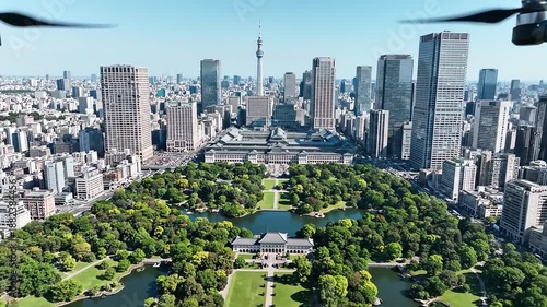 A high-angle aerial shot captures the vibrant green imperial palace east garden, dramatically contrasting the lush park and moats with the dense modern skyscrapers of central tokyo, japan.