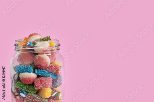 Cropped view of the upper half of a glass jar filled with colorful sugared gummy candies on the left side of a bright pink background with copy space on the right. Close-up, isolated, front view