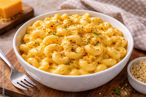 Mac and cheese served in a white bowl on a wooden board. Homemade comfort food with creamy cheese sauce, garnished with spices and herbs, concept of cozy meal and hearty lunch.