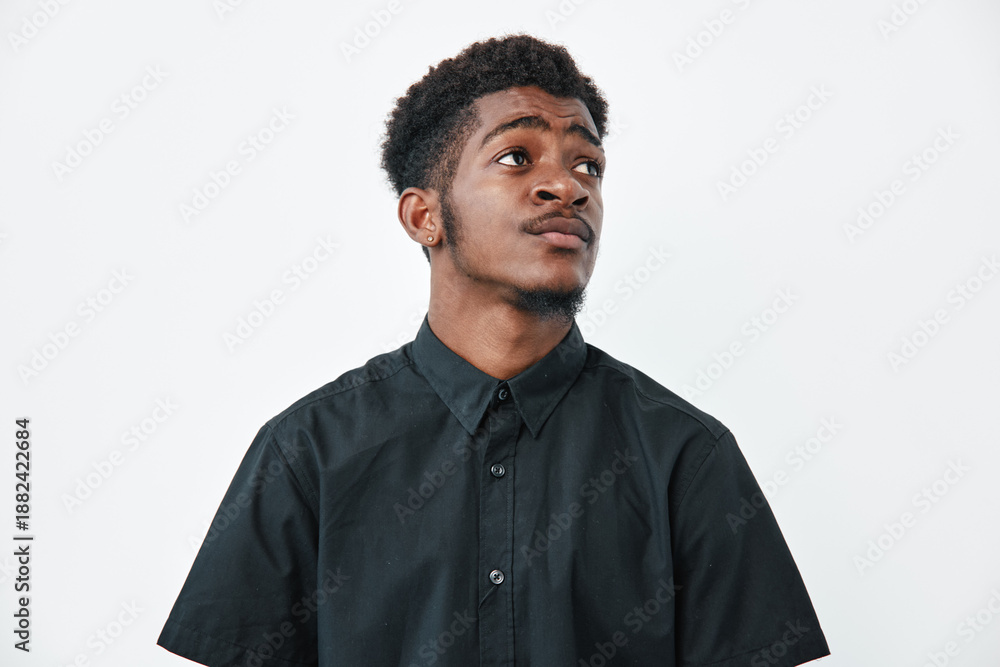 Obraz premium Young black man with dark skin wearing black shirt looking up thoughtfully on white background. Portrait of African ethnicity male with short curly hair and casual style.