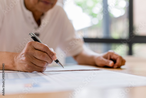 Senior man writing on document at home, close-up of hand holding pen. Retirement planning, financial paperwork, insurance form, legal document, senior lifestyle and careful decision making concept.