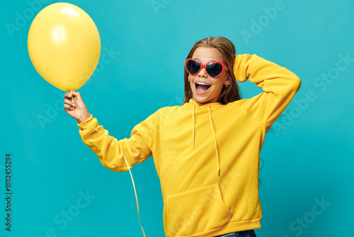 Bilde på lerret Girl in yellow hoodie and red heart sunglasses holding yellow balloon on blue background with joyful expression smiling for fun and celebration event or party moment