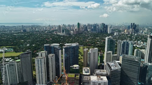 Wide aerial perspective of Bonifacio Global City featuring modern high-rise towers, green parks, and distant Manila Bay coastline under bright daylight.