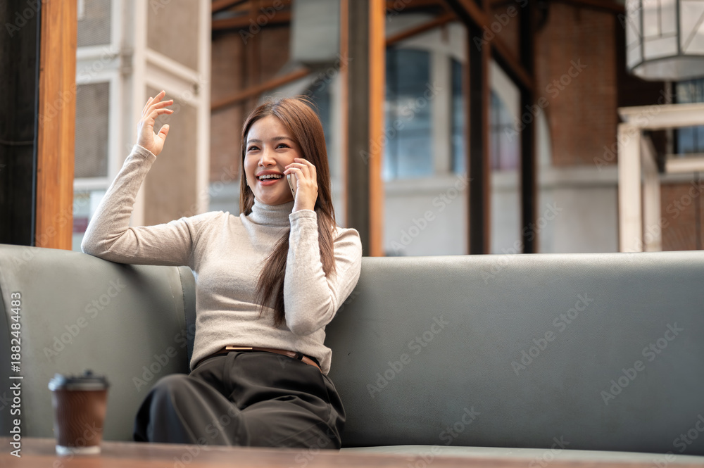 Fototapeta premium Pretty asian woman talking on phone with one hand raised while sitting on sofa in coffeehouse cafe.
