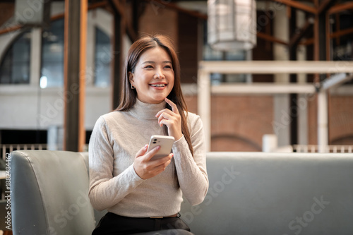 Pretty asian woman holding phone thinking with finger under chin while sitting on sofa couch in cafe