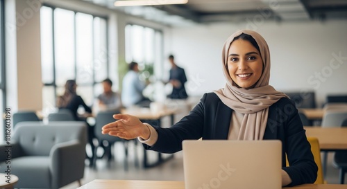 A welcoming Muslim woman in a hijab, seated at a desk with a laptop, extends her hand in a gesture of invitation or presentation in a bright, modern office, business concept, modern, welcoming