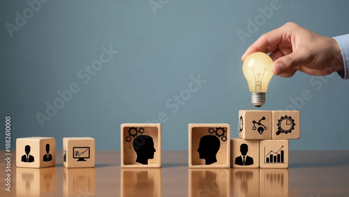 Hand placing a lightbulb on wooden blocks with business icons on a reflective desk with a blue background.