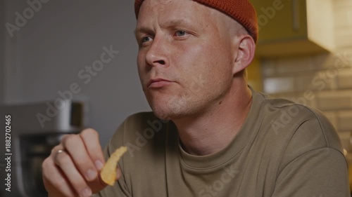Medium side-angle shot of a man chewing a potato chip while sitting at a kitchen table. Camera positioned slightly to the side, warm indoor lighting, relaxed home snack moment. High quality 4k footage
