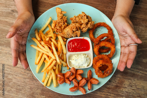 Foto Top view of a hand holding a mix snack platter, which includes chicken wings, onion rings, French fries, sausages, as well as chili sauce and mayonnaise as condiments