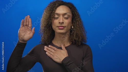 Woman raising her palm and placing her other hand on chest in a blue studio setting; pledge sincerity.