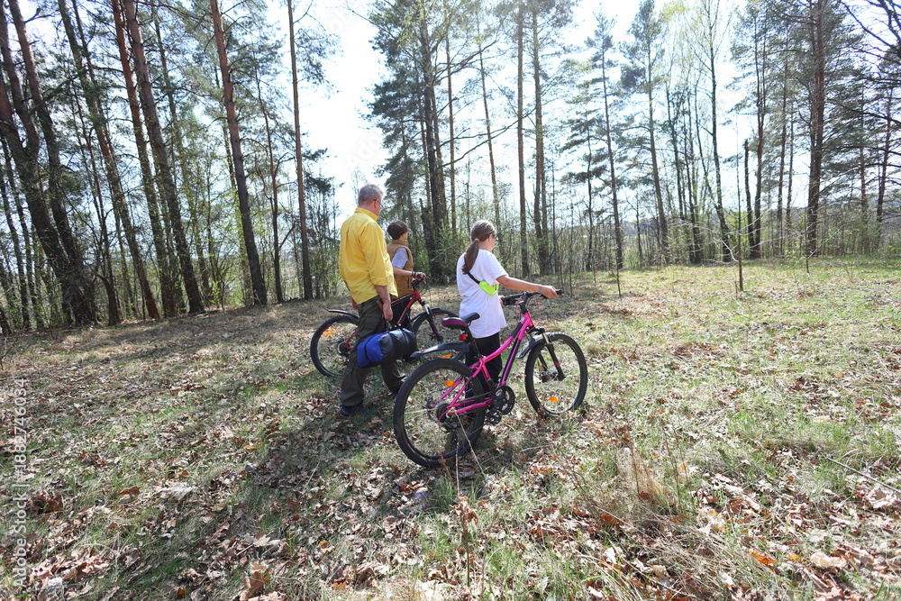 Fototapeta premium A father and two kids walk with bikes through a sun-dappled forest, heading toward adventure. A peaceful moment of family bonding and outdoor exploration in nature.
