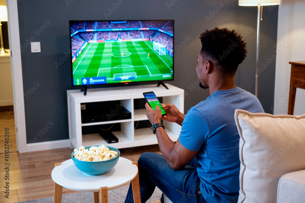Fototapeta premium Black young adult man sitting on couch watching soccer match on television while holding smartphone in living room, bowl of popcorn on table in foreground, focused on screen