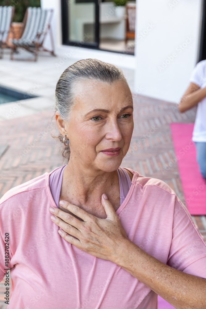 Fototapeta premium Senior woman sitting on brick patio by pool wearing pink top, sports bra, yoga mat right