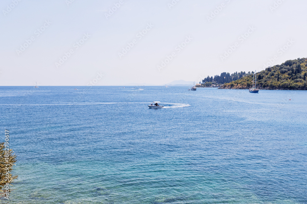 Obraz premium Speedboat cruising across turquoise sea with sailboats and green coastline in distance