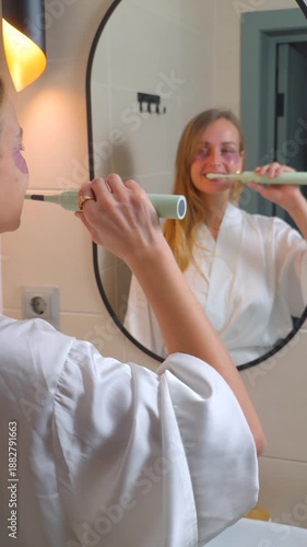 Woman brushing teeth with electric toothbrush while looking in bathroom mirror