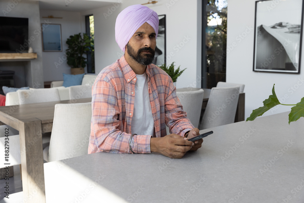 Naklejka premium Indian man wearing lavender turban sitting at gray kitchen island holding smartphone in dining area