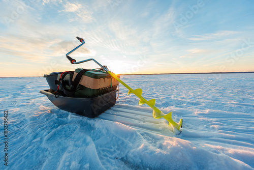 фотографія A hand auger with yellow blades rests by a black gear sled on a wide frozen lake at sunrise