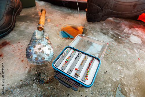 Close view shows jigs, spoons, skimmer, and winter boots on rough ice. A freshly drilled hole with slush sits center as an angler selects lures in cold daylight.
