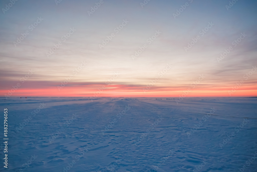 Fototapeta premium A wide frozen expanse shows faint tracks and distant huts for ice fishing. Low sun casts long shadows toward the horizon. Soft light and minimalist composition suggest isolation.