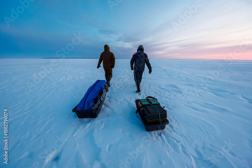 Obraz na plátně Two bundled anglers walk on open ice at dawn, pulling sleds with a shelter bag and tackle boxes