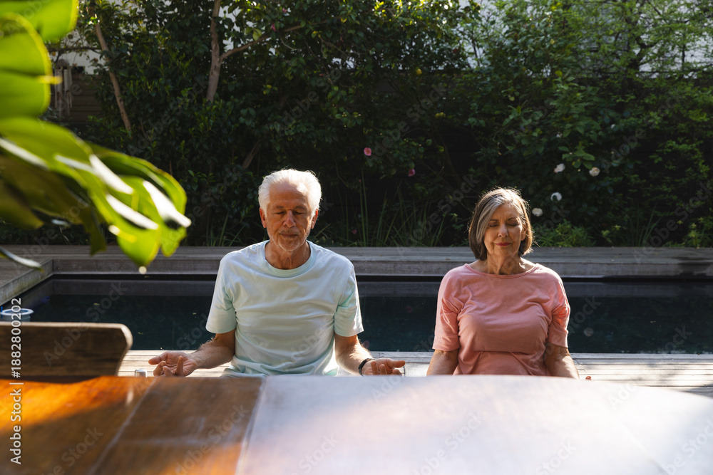 Fototapeta premium Senior couple meditating cross-legged on wooden pool deck by pool in backyard near table and shrubs