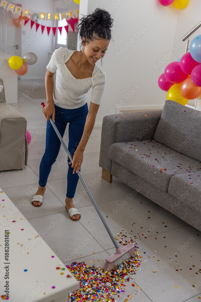 Obraz premium Female teenager sweeping confetti from tiled living room floor with broom near balloons