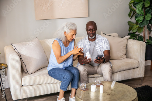 Diverse senior couple reviewing medication instructions on smartphone and pill bottles on home sofa