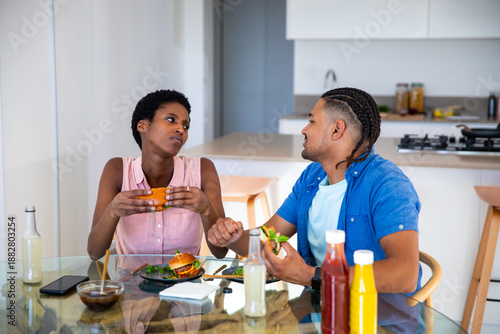 African American and Hispanic couple eating burgers at kitchen table with salad, condiment bottles