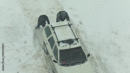 Two people push white car stuck in heavy snow on unplowed road. Winter weather creates hazardous conditions for transport as people work to free vehicle.
