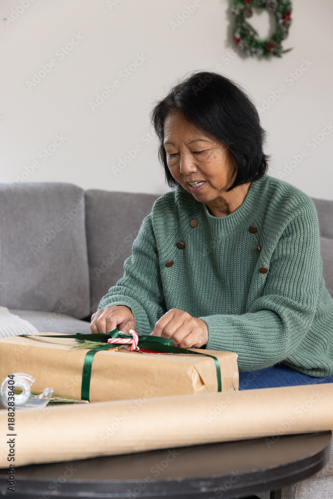 Obraz premium Senior Asian woman wrapping gift box with green ribbon on table in living room, copy space