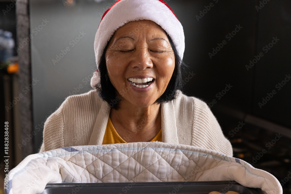 Fototapeta premium Senior Asian woman leaning over baby bassinet with quilted blanket in living room wearing Santa hat