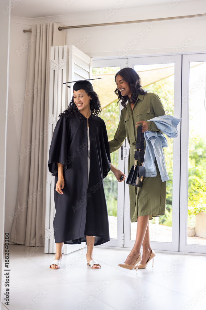Fototapeta premium Mother guiding teenage daughter in home glass doorway holding graduation cap, gown