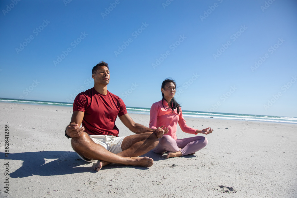 Obraz premium Diverse couple practicing meditation sitting cross-legged on oceanfront beach under clear sky